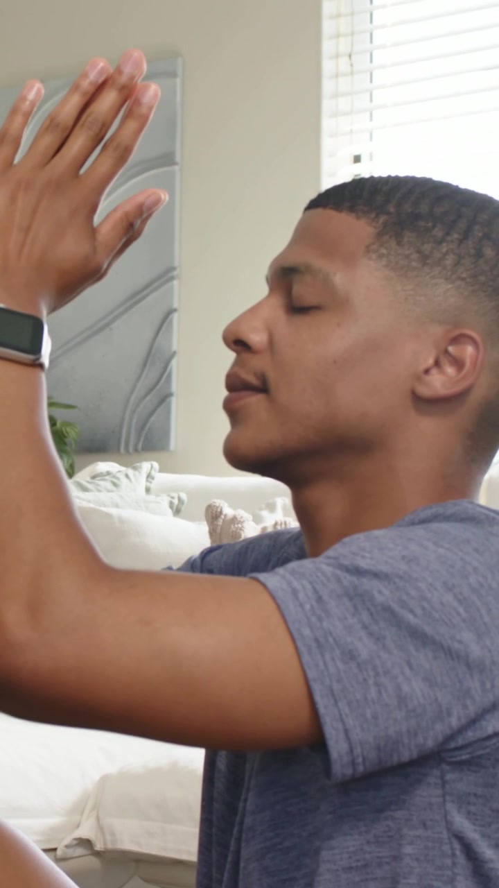Video verticale: Dopo aver alzato le braccia, uomo con maglietta blu che giunge i palmi, si calma sul divano di casa, con orologio.