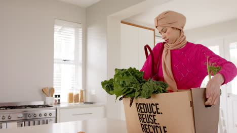mujer biracial en hijab con compras de comestibles en la cocina en casa con espacio de copia, cámara lenta