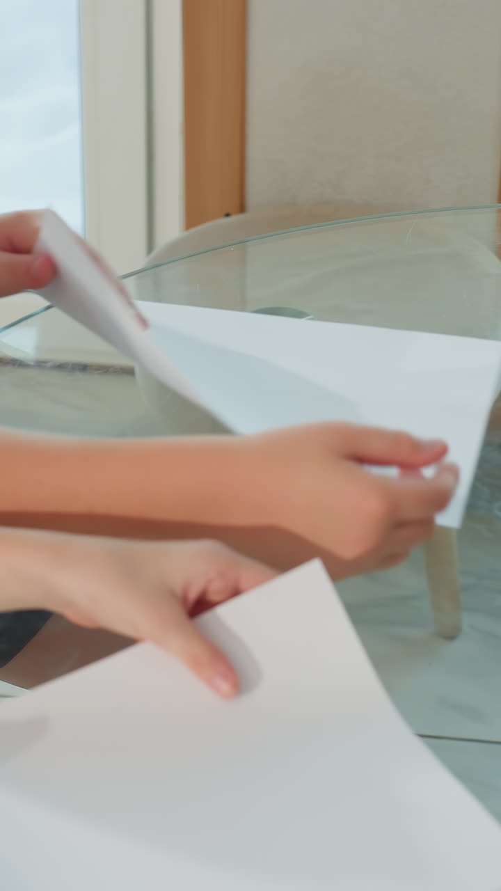 Niños doblando papel para hacer aviones en una mesa de cristal, barra de pegamento en el borde, manos intercambiando hojas, luz brillante del día entrando por la ventana, fondo nevado en el exterior, energía juguetona y llena de risas, trabajo en equipo y aprendizaje