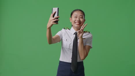 아시아 여성 조종사가 스마트폰을 사용하여 녹색 화면 배경 스튜디오에 서서 사진을 찍습니다.
