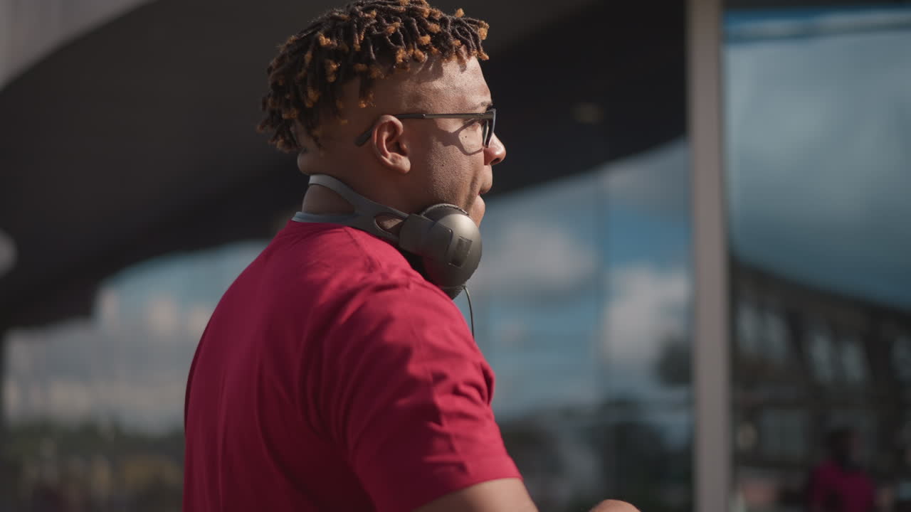Outdoor black DJ mixing on laptop, cueing tracks and adjusting controller near glass architecture, headphones around neck, concentrated expression, reflective windows and daytime event energy