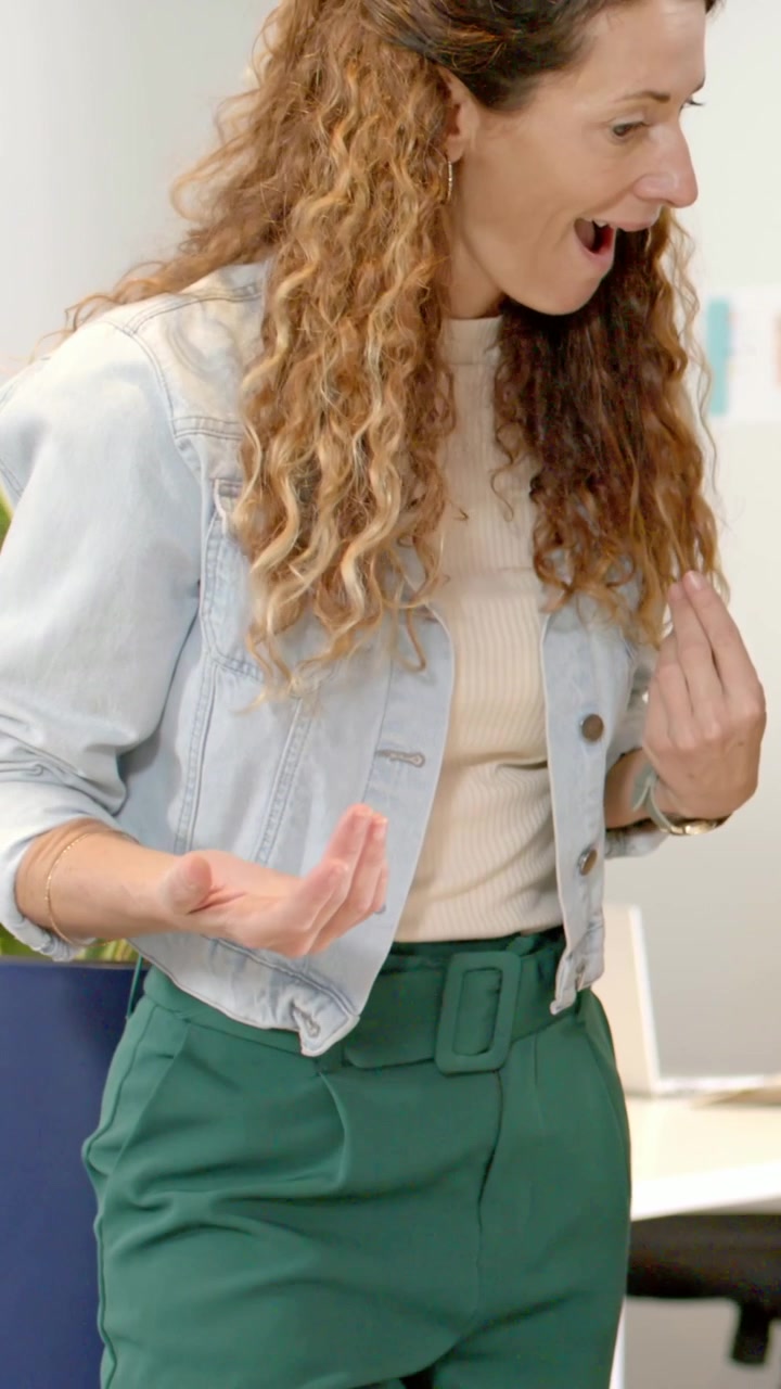 Video verticale: Donna in jeans che si sporge in avanti e gesticola, spiegando alla scrivania con telefono e taccuino.