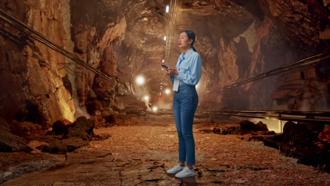 full body side view of asian female worker with her phone in underground mine tunnel, she observes by looking around before she come to concentrating with her smartphone