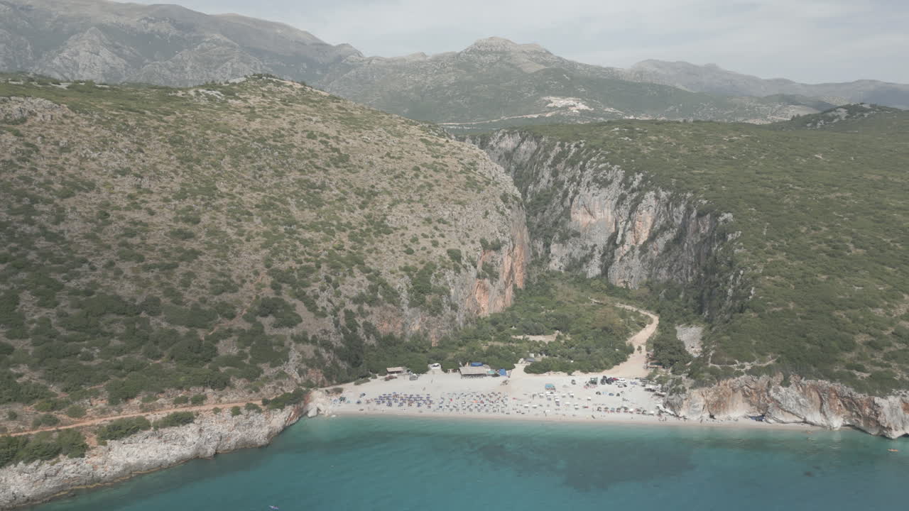 Drone shot over Gjipe beach Albania with tourists on the beach and clear blue water near the mountains and the sea on a sunny and bright day and green nature around with sun beds towels LOG