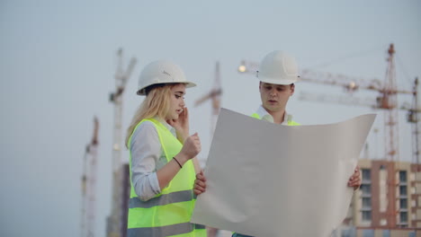 workers with drawings at the construction site. two workers man and woman in protective harhats working with drawings at the construction site outdoors.