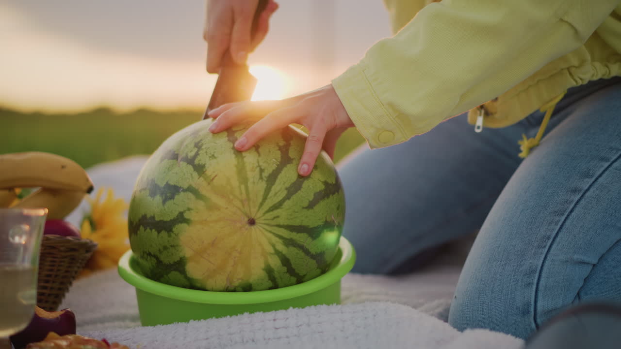Persona cortando sandía a mano al atardecer; viste una chaqueta amarilla y vaqueros mientras prepara un aperitivo veraniego sobre una manta. Se ve un soporte de melón verde y una cesta de mimbre. Resplandor de la hora dorada, pradera cubierta de hierba.