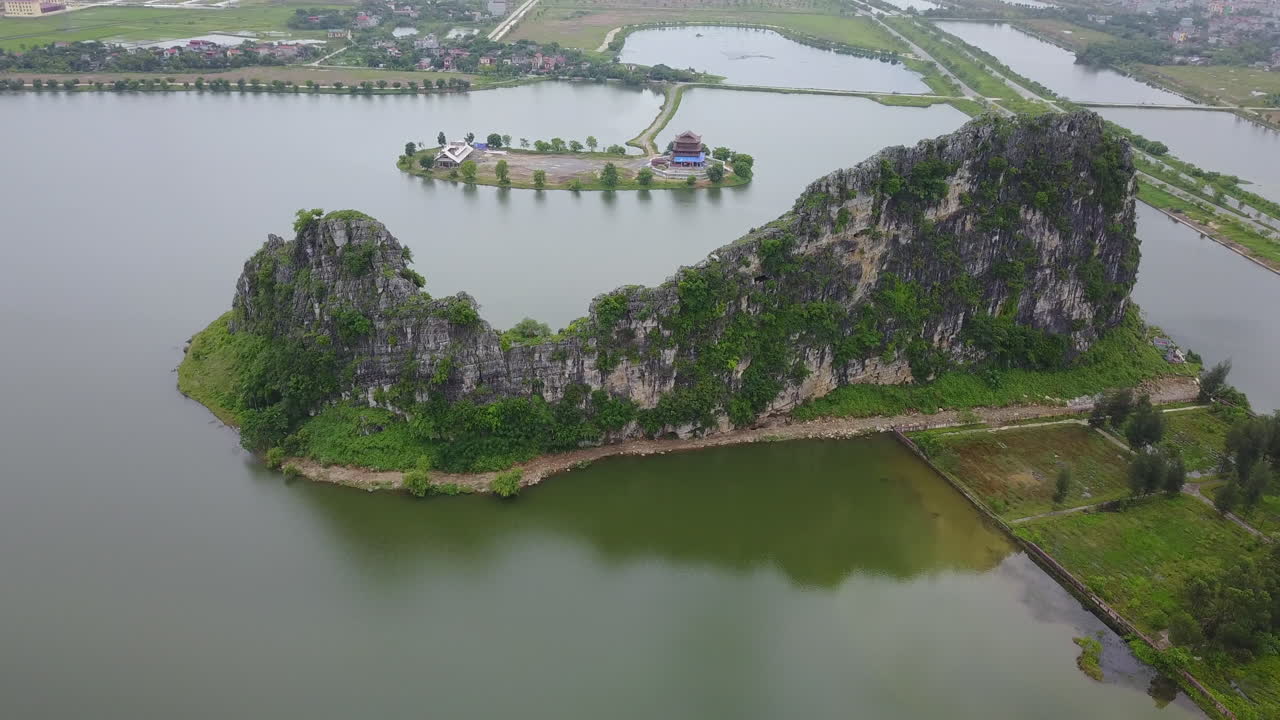 Experience a zoom-out view near a unique rock formation situated beside the Dinh Thanh Cao Son temple in Trang An, Ninh Binh, Vietnam, revealing the temple's setting amidst the serene waters