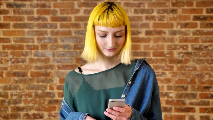 Giovane donna affascinante con i capelli gialli che digita al telefono e sorride, in piedi vicino a un muro di mattoni sullo sfondo, persona insolita, straordinaria.