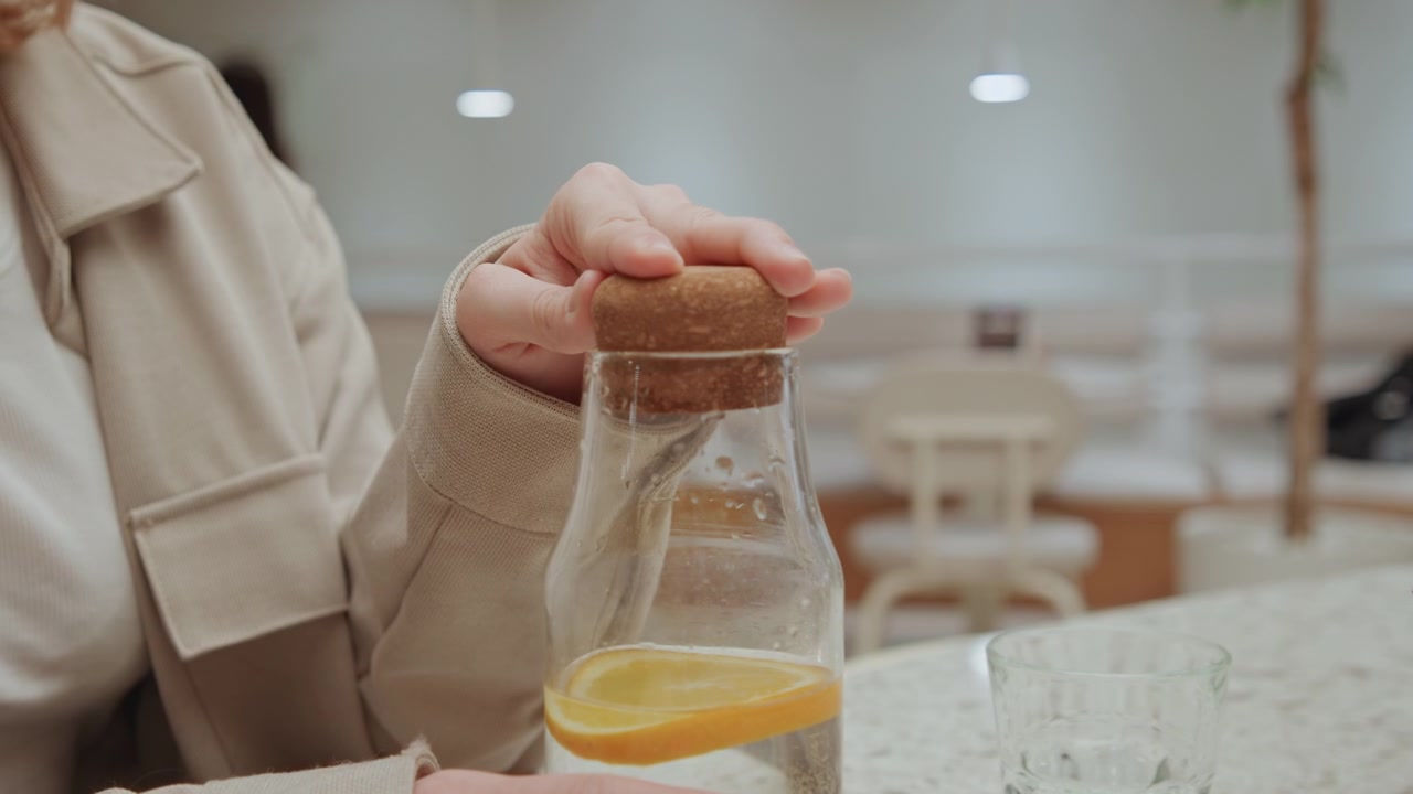 Primo piano di una donna con un cappotto beige che tiene e appoggia un oggetto sopra dell'acqua con una fetta di limone. Bicchieri e fiori rosa sono visibili sul piano del tavolo. Ambiente interno di un caffè con illuminazione soffusa.