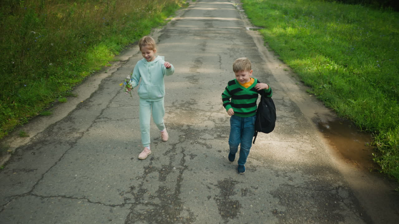 Smiling girl holding flowers and cheerful boy with bag share a joyful moment walking side by side on quiet paved path through lush forest, enjoying conversation and laughter under sunlight