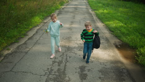 smiling girl holding flowers and cheerful boy with bag share a joyful moment walking side by side on quiet paved path through lush forest, enjoying conversation and laughter under sunlight