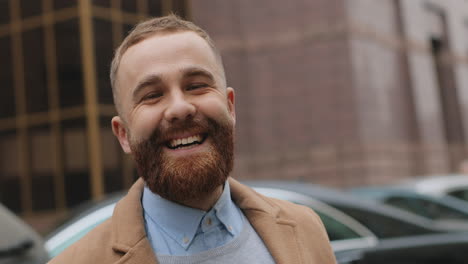 Close-up-view-of-caucasian-young-businessman-with-a-beard-wearing-elegant-clothes-looking-and-smiling-to-the-camera-in-the-street-in-autumn