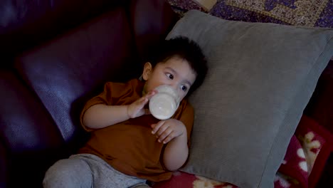 adorable 18-month-old baby boy sits leisurely on a home sofa, holding a bottle of milk and drinking from it, bathed in the warm glow of tv screen light reflections