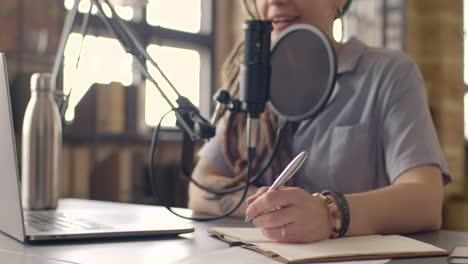 vista de cerca de una mujer grabando un podcast y tomando notas sentada en una mesa con un micrófono y una computadora portátil