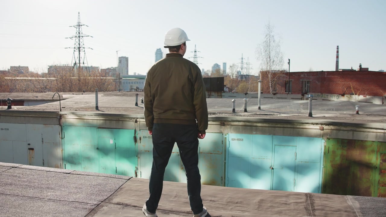 Trabajador inmóvil en un tejado de asfalto cerca de un parapeto, visto desde atrás con luz diurna tenue, con casco de seguridad, chaqueta, pantalones oscuros y zapatillas, observando la infraestructura de la ciudad y el tejado