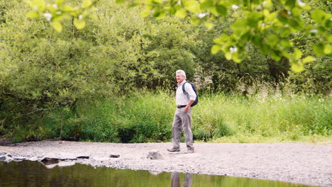 Slow-Motion-Shot-Of-Senior-Man-Using-Stepping-Stones-To-Cross-River-Whilst-Hiking-In-UK-Lake-District