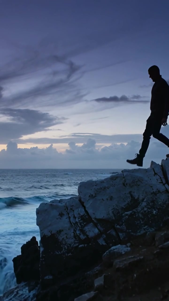 Video verticale: Uomo che cammina con giacca e stivali su una scogliera al tramonto, evitando gli spruzzi delle onde che si infrangono, con spazio per il testo.