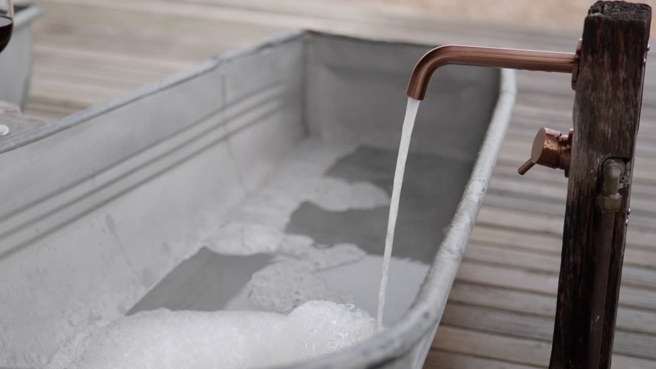 Static handheld close-up of copper tap streaming water into foamy metal bathtub with sharp focus on fixture and soft bokeh background