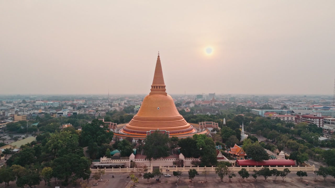 Un volo fluido di drone avanza verso un'enorme stupa dorata che si erge sopra il centro urbano, mentre un maestoso monumento buddista domina lo skyline della città in Thailandia sotto un cielo grigio e velato con il sole mattutino splendente