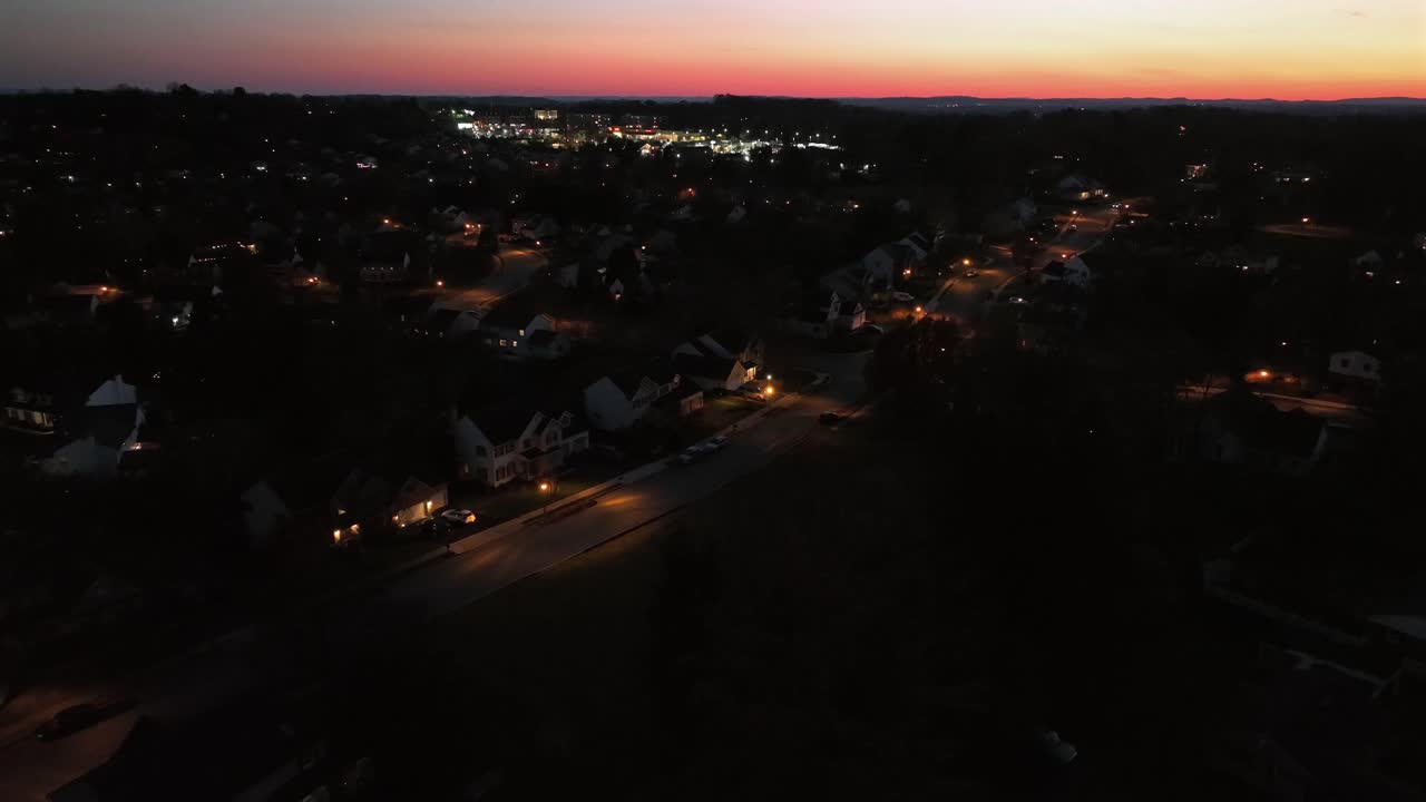 Aerial dusk view of glowing American city neighborhood, warm streetlights lining residential blocks while distant urban center shines under orange horizon fading into deep evening sky. Wide shot