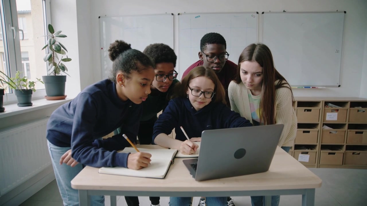 Reacting to laptop prompt, group students in casual clothes taking notes for project at class desk