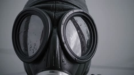 raindrops steadily falling on black gas mask lens, creating haunting visual symbolizing survival, protection, and potential environmental threat against stark white background