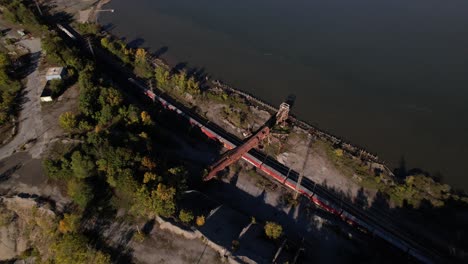 an aerial view over train tracks as a long cargo train goes by, on the edge of the hudson river in upstate ny