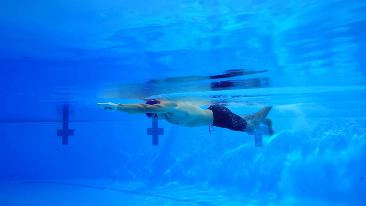 Persona che nuota sott'acqua attraverso una scena di piscina blu con movimento fluido in avanti, bolle visibili e distorsione dell'acqua, riflessi superficiali illuminati, composizione calma e un ambiente acquatico limpido.