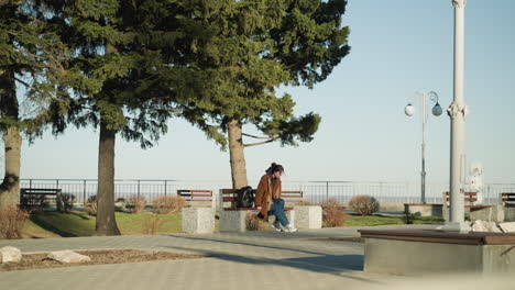 young woman wearing a brown coat, blue jeans, and white shoes sits alone on a bench in an urban park, her black backpack beside her. she appears contemplative, with a isolated atmosphere surrounding