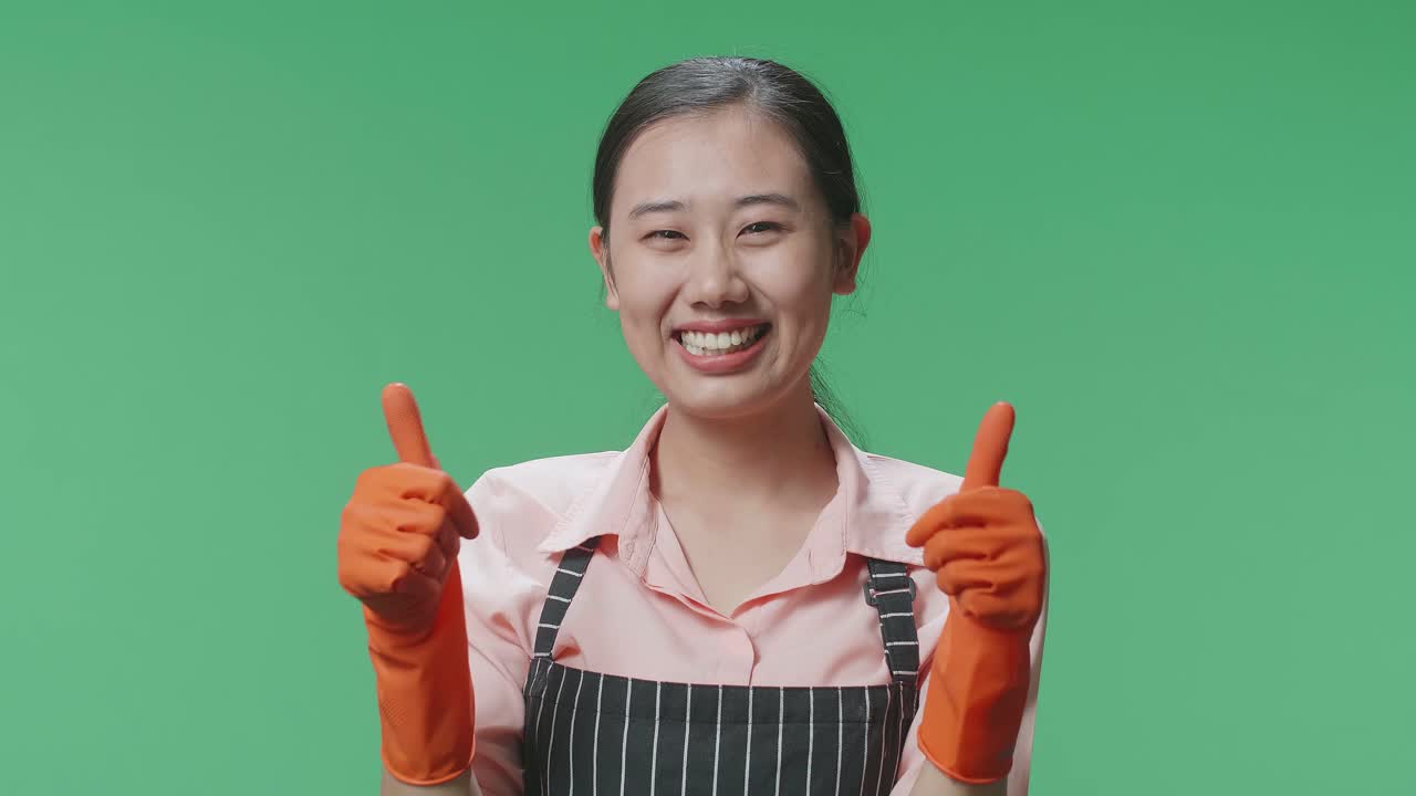 グリーンスクリーンスタジオに立っている間アプロンを着て笑顔で親指を上げているアジア人女性家政婦のクローズアップ