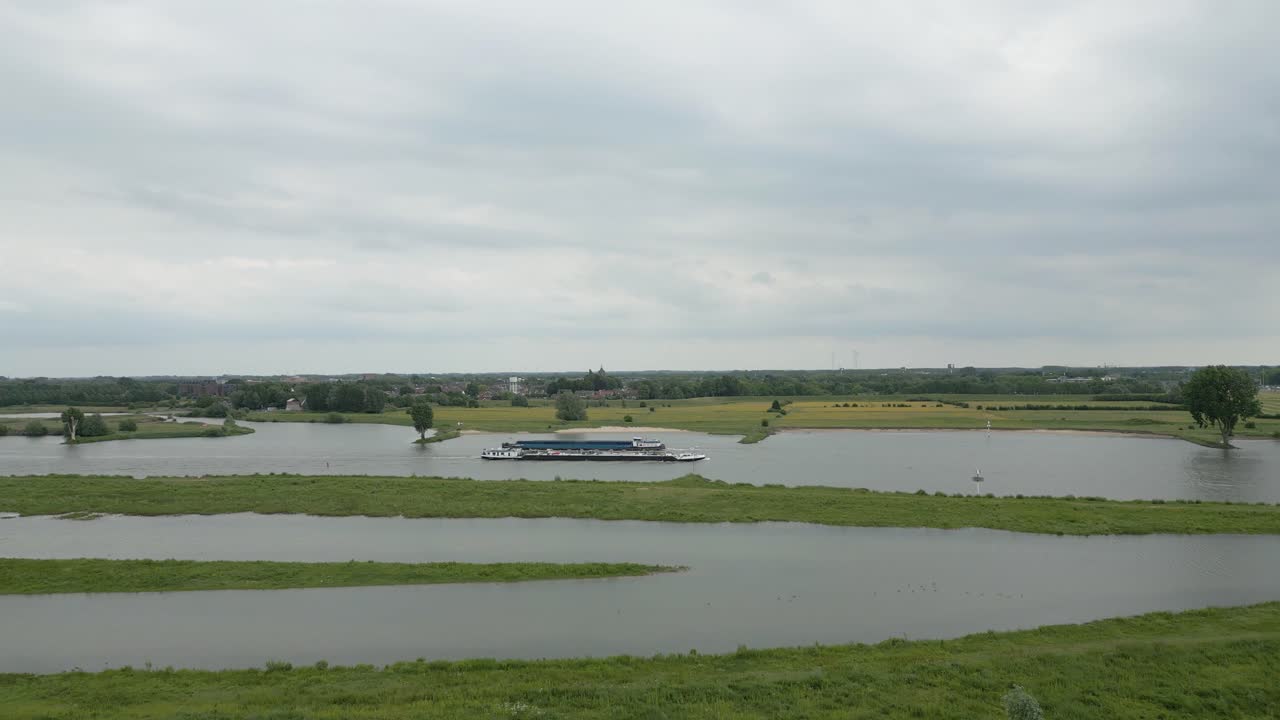 Wide drone view of green wetlands with a cargo ship gliding along the river under a cloudy sky.