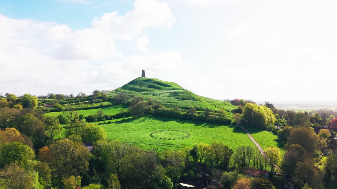 Birleşik Krallık'taki ikonik tepeyi ve kuleyi gösteren, Somerset İngiltere'deki ağaçlar ve tarlalarla yeşil kırsalın üzerinde yükselen Glastonbury Tor'un geniş havadan görünümü