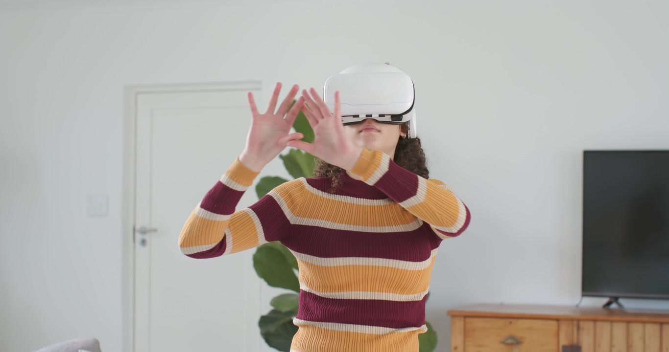 Giovane donna afroamericana con maglietta a righe, che indossa un visore e sta avviando, allungando la mano e usando il menu VR vicino alla TV