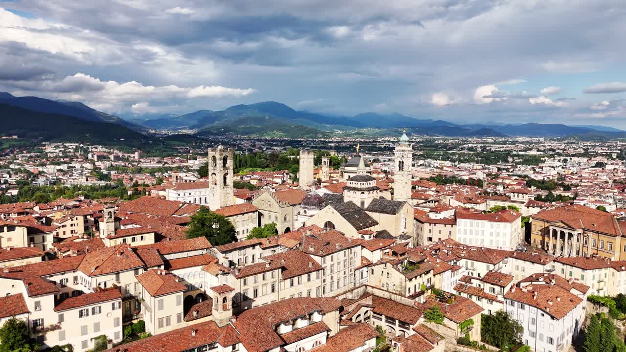 Aerial panorama of Bergamo Italy featuring dense terracotta rooftops, historic towers, stone buildings and distant mountains, with sunlight showing city layout and surrounding valley under bright sky