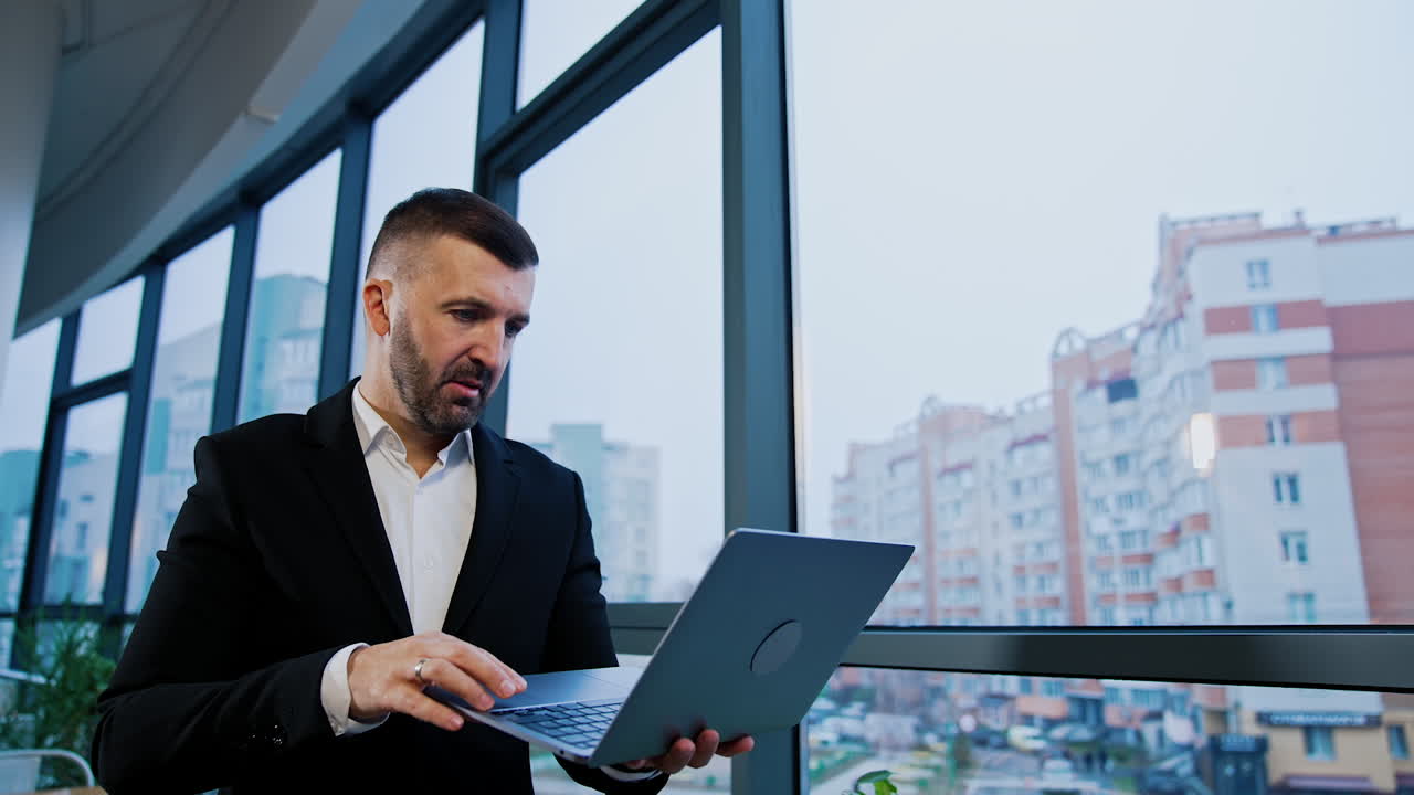 Thoughtful businessman stands holding laptop in hands. Male boss types on computer waving head disapprovingly. Low angle view.