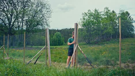 a man works on securing a wooden fence post in a grassy field surrounded by trees - static shot