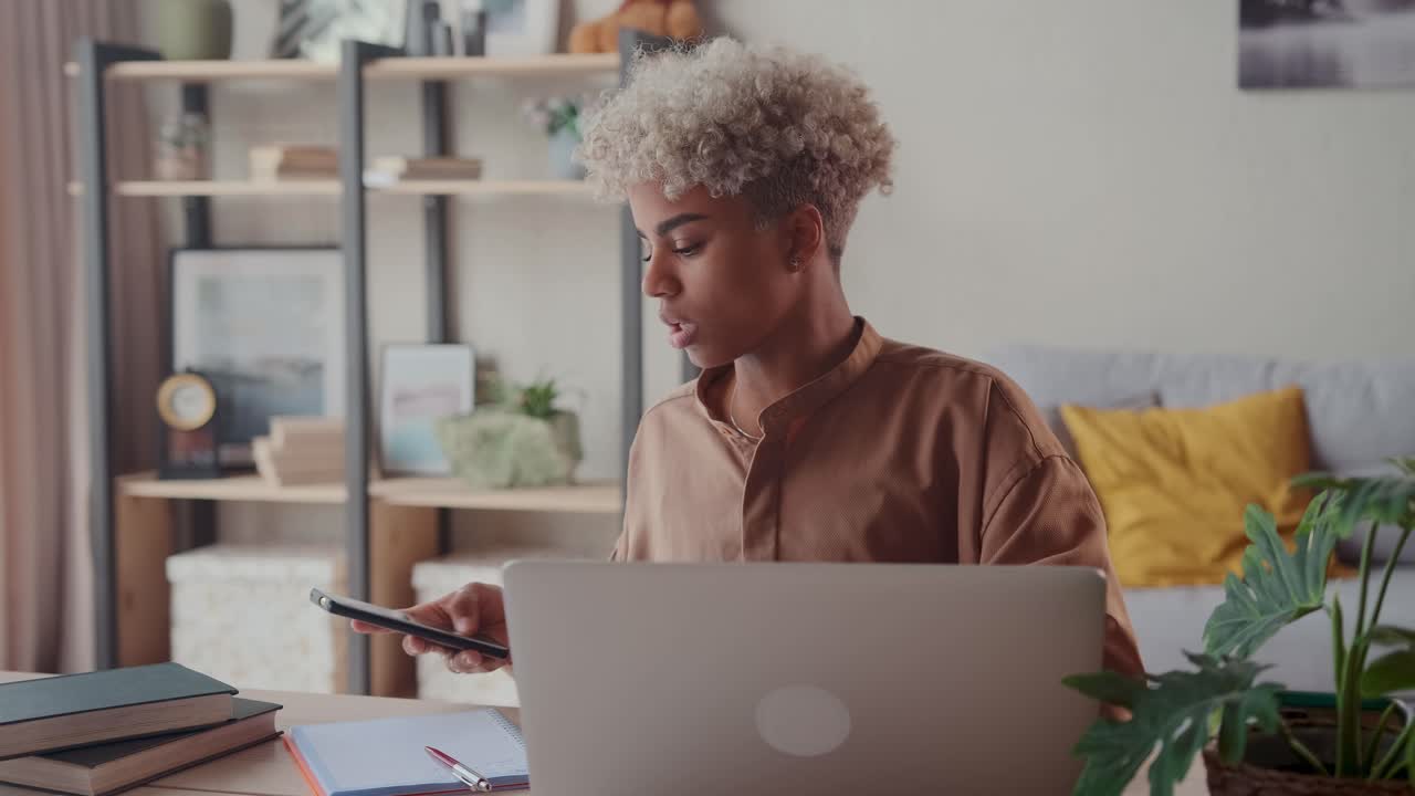 Young african american woman student holding phone sits at table with laptop