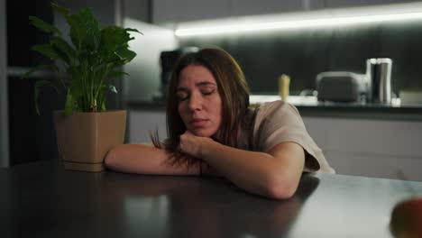 A-sad-and-tired-brunette-girl-in-a-beige-T-shirt-sits-leaning-on-a-black-dining-table-near-a-green-houseplant-in-a-modern-kitchen-in-the-evening