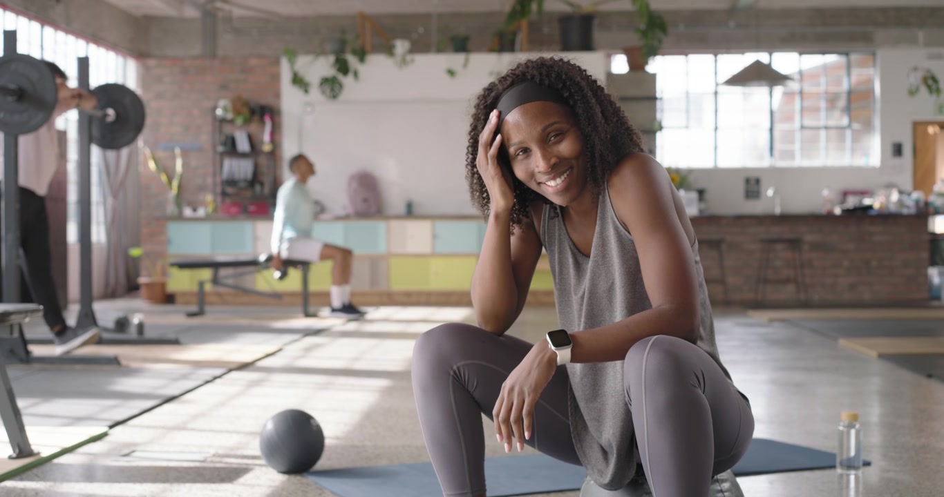 Donna afroamericana in una palestra loft, in recupero su una palla, che lancia un'occhiata a un sollevatore di bilancieri, rivolta verso la telecamera.