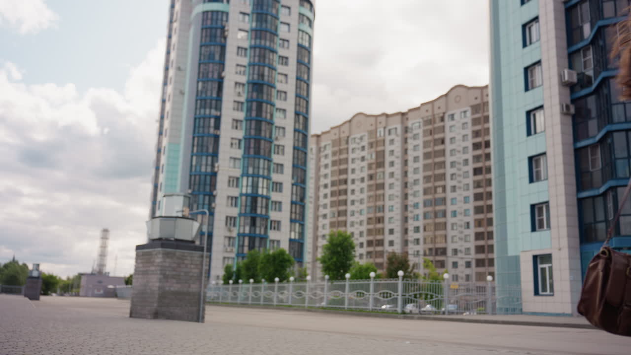 Camera shows astonishing view of young woman cat walking in cozy urban area, She moves with relaxed confidence holding parasol, framed by modern towers, wide ground perspective under clouded sky