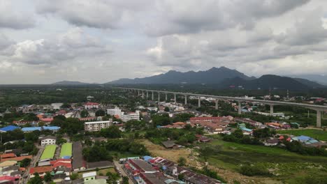 Imágenes-Aéreas-Deslizándose-Hacia-La-Izquierda-De-Muak-Lek-Y-Montañas-En-El-Horizonte,-Un-Ferrocarril-Elevado-De-Alta-Velocidad-A-Través-De-La-Ciudad-Durante-Un-Día-Nublado-En-Saraburi,-Tailandia