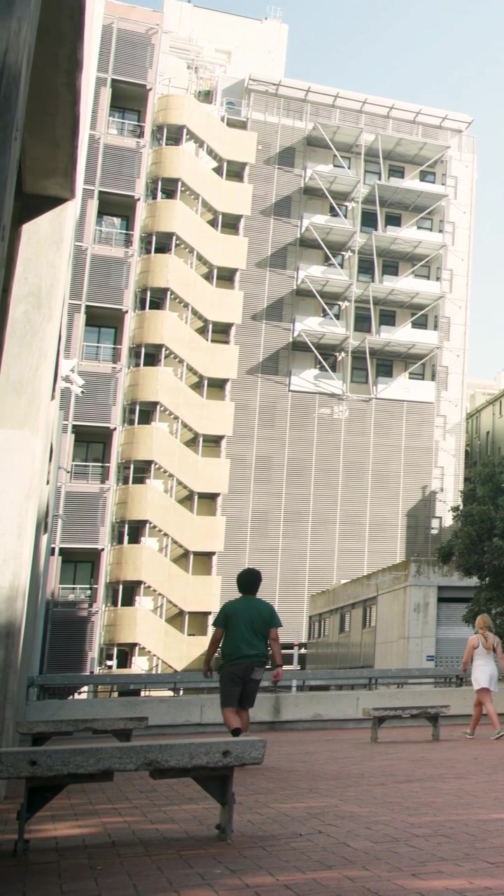 Video vertical: Hombre con camiseta verde y pantalones cortos oscuros entrando en cuadro, caminando hacia una barandilla en la plaza, con un banco.