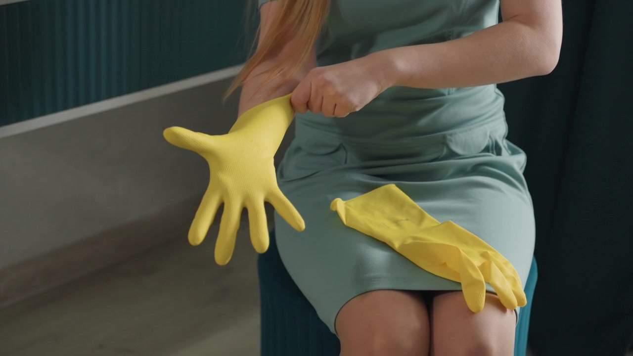 Proprietaria di casa che si sta mettendo i guanti gialli, seduta su uno sgabello in un interno accogliente, sotto una luce naturale soffusa, con un vestito verde, le mani che sistemano i guanti di gomma, con movimenti lenti e deliberati, in preparazione alle pulizie mattutine, in un ambiente domestico ordinato.