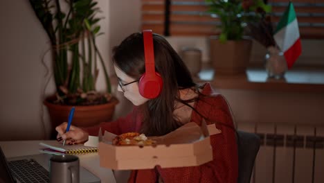 Side-view-of-a-confident-brunette-girl-in-red-wireless-headphones-studying-foreign-languages-near-the-flag-of-Italy-using-online-lessons-on-a-laptop-and-taking-notes-in-her-notes-while-eating-pizza-in-a-modern-apartment-in-the-kitchen