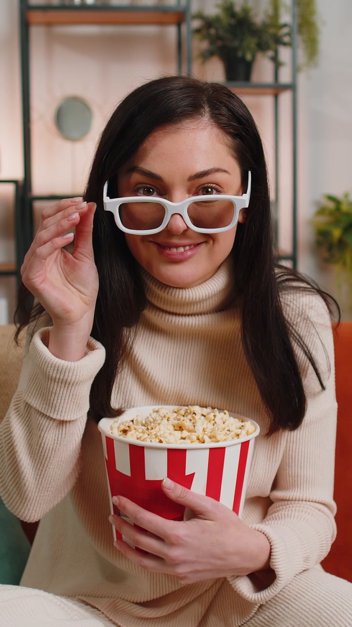 Donna seduta sul divano che mangia popcorn e guarda un'interessante serie TV o una partita sportiva online a casa