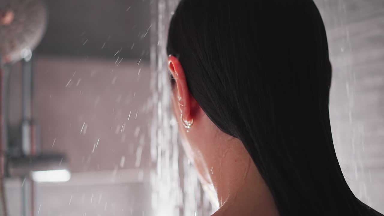 황금 수입을 가진 여자가 샤워실에 서 있습니다. 여자는 뜨거운 물을 즐기고 샤워에서 몸 아래로 흐릅니다. 여자는 더운 물 아래에서 화장실 위생 절차를 수행합니다.