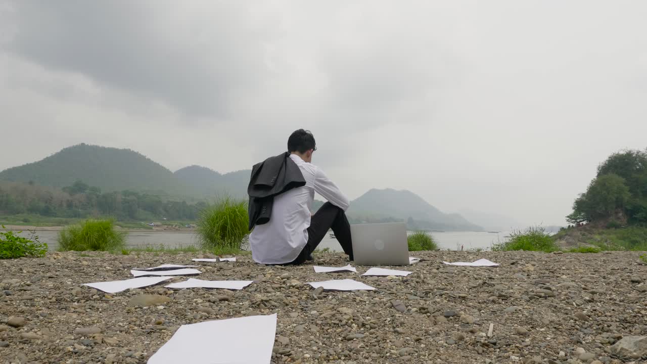 Businessman relaxing outdoors by the river with laptop and documents