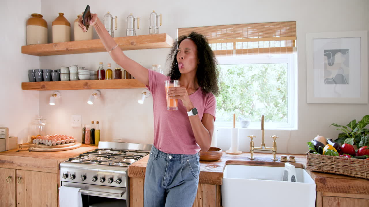 Gustando succo fresco in una cucina accogliente, una donna sorridente che tiene in mano lo smartphone, a casa.