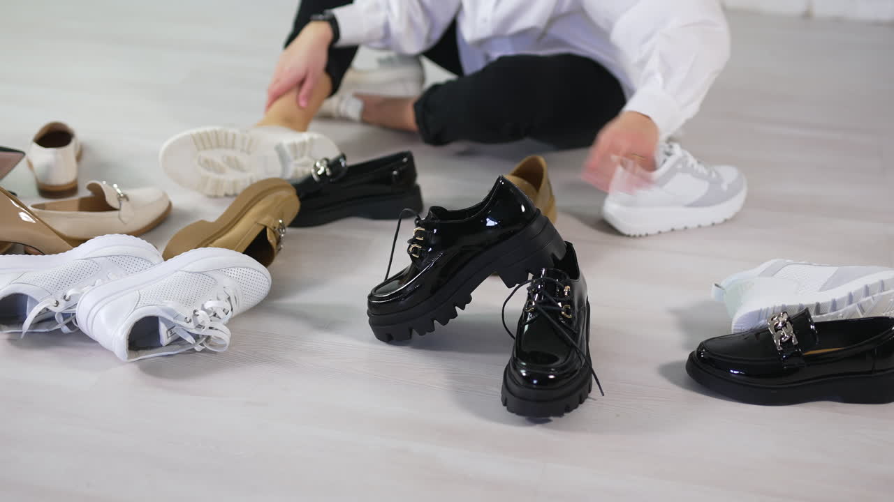 Woman sits on the floor surrounded by new fashionable footwear. Lady rejects all shoe variants choosing the black lacquered ones.