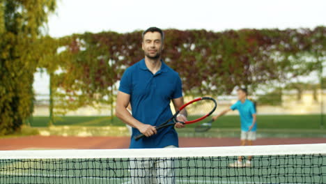 Portrait-Of-A-Handsome-Man-Coming-Closer-To-Camera-With-Racket-In-Hand,-Leaning-On-Net-And-Smiling-At-The-Camera-While-Spending-Time-With-His-Family-On-A-Tennis-Court